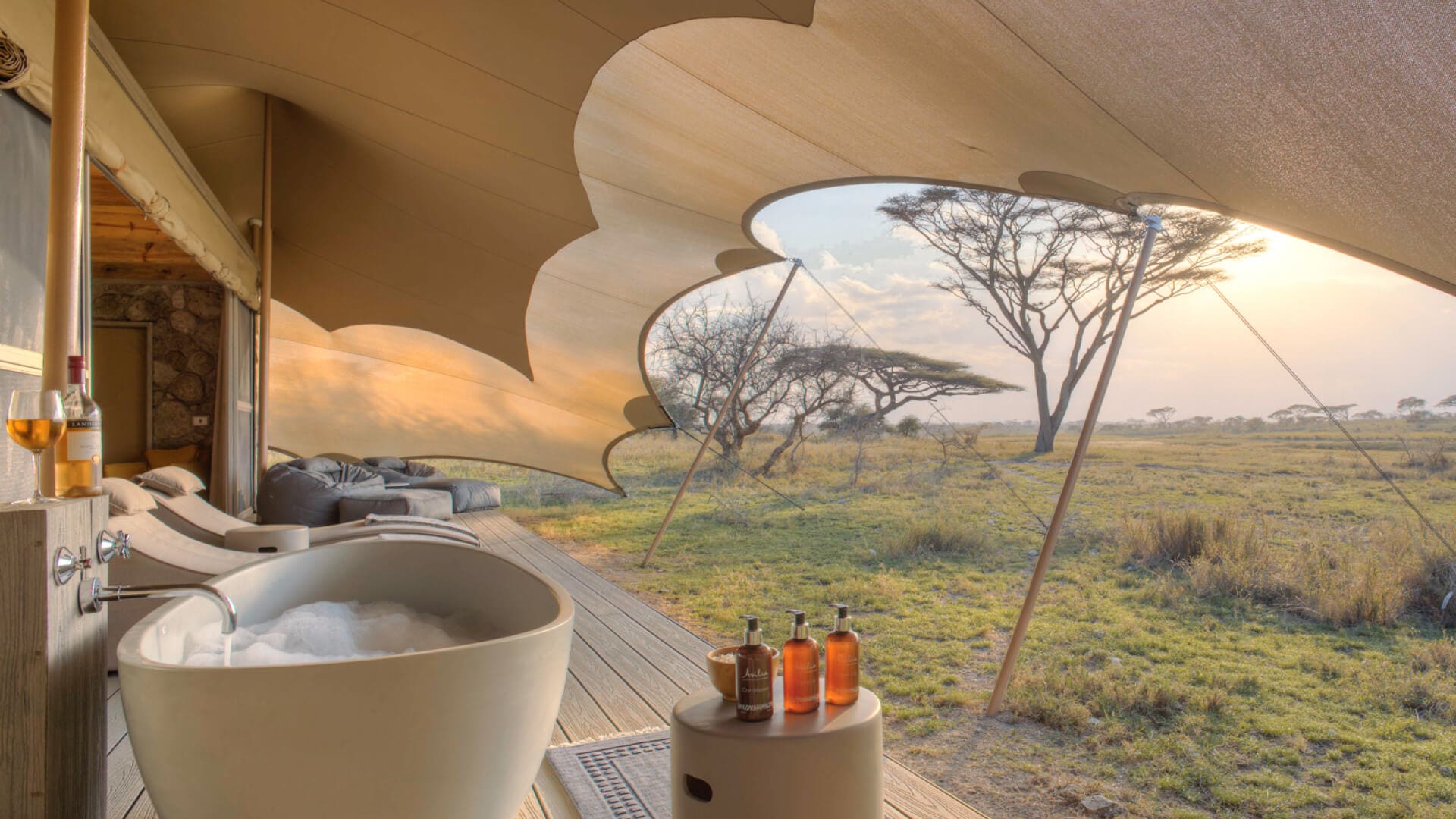 Namiri Plains, Serengeti National Park, Bath on the deck with bubbles and wine glass and bottle, amenity bottles and bath salts, loungers and bean bags